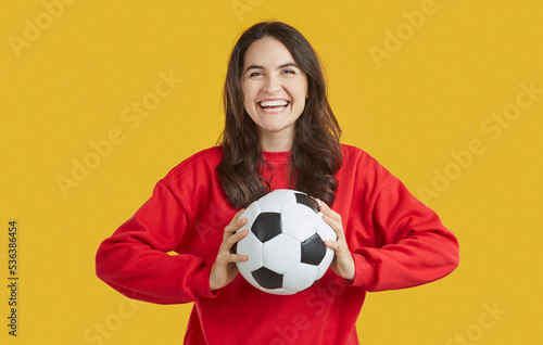 Portrait of cheerful positive young woman who is having fun catching soccer ball thrown to her. Young woman with traditional black and white soccer ball in hands laughing isolated on orange background © Studio Romantic