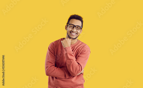 Studio portrait of happy cheerful college or university student. Handsome young black man in sweatshirt and glasses holding hand on chin, smiling and looking at camera isolated on yellow background