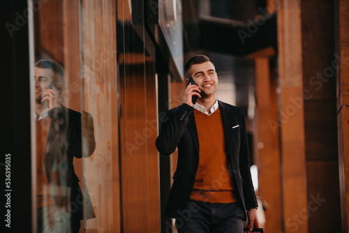 Going to airport terminal. Confident businessman traveler walking on city streets and pulling his suitcase drinking coffee and speaking on smartphone