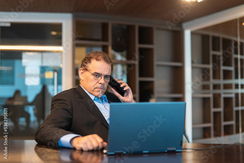 Portrait of mature businessman using laptop computer and mobile phone in office