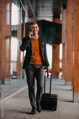 Going to airport terminal. Confident businessman traveler walking on city streets and pulling his suitcase drinking coffee and speaking on smartphone