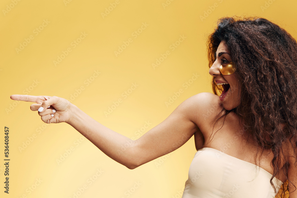Obraz premium Amazed happy pretty Latin female with gold hydrogel patches under eye point finger look aside posing isolated over yellow background. Cosmetic product ad Natural beauty concept Studio profile