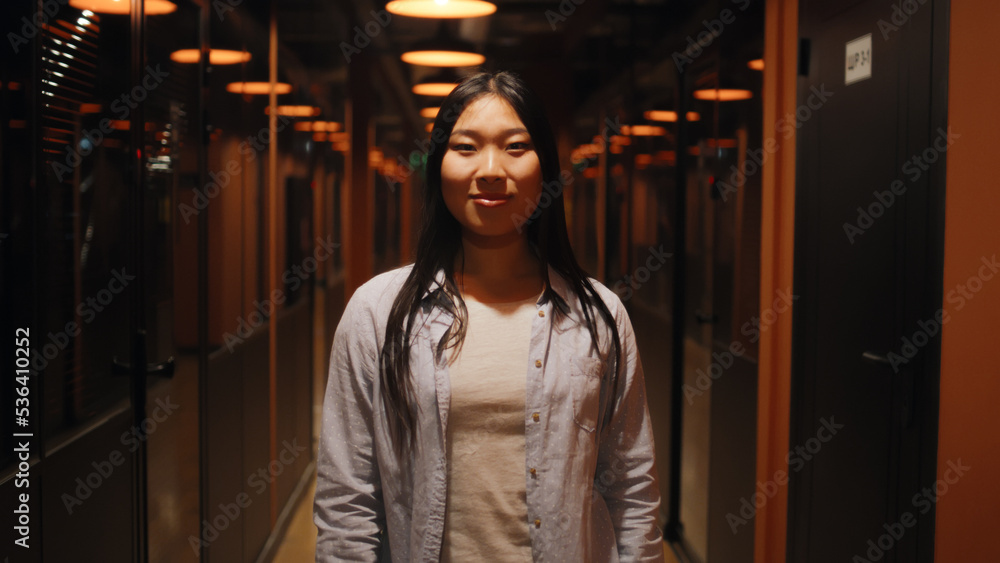 Happy and successful asian woman standing in office hallway, smiling, looking at camera. Motivated female office worker in corridor of marketing company modern business center
