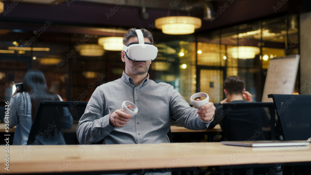 Male office worker wearing VR headset and using wireless controllers, gesturing, watching data in virtual reality. Work in office of hi-tech company near coworkers. Cyberspace digital technology Male office worker wearing VR headset and using wireless controllers, gesturing, watching data in virtual reality. Work in office of hi-tech company near coworkers. Cyberspace digital technology