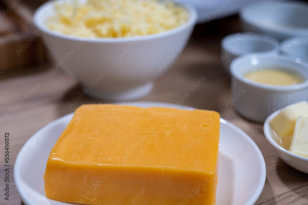 A piece of cheddar cheese on a white plate in the kitchen on a wooden table and white bowls with food ingredients on a blurred background. food background. Healthy food advertisement, poster for the k
