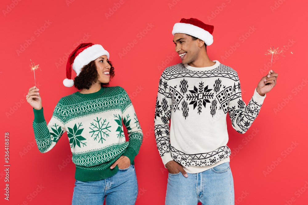 Fototapeta premium young african american couple in santa hats holding sparklers and smiling at each other isolated on red