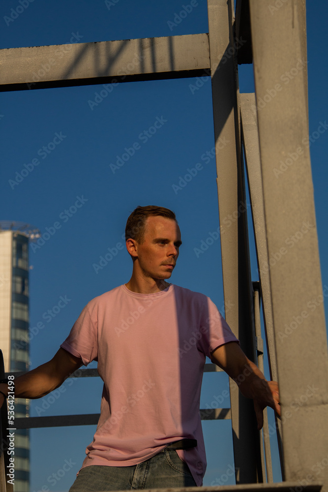 A handsome guy in denim shorts and a pink t-shirt poses beautifully in the city