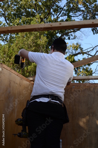 Jewish boy wearing white shirt, kippah, and black pants building sukkah with wooden boards and drills for sukkot/sukkos holiday photo
