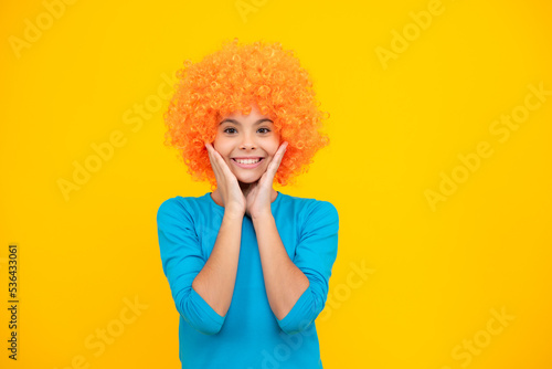 Birthday kids party. Funny kid in curly clown wig isolated on yellow background. Happy teenager portrait. Smiling girl.