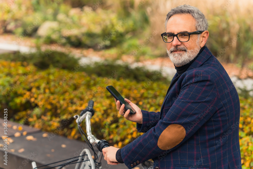 Technology in the life of elderly people. Handsome caucasian gray-haired bearded man in black-frame glasses holding his smartphone and holding bicycle wheel. High quality photo Technology in the life of elderly people. Handsome caucasian gray-haired bearded man in black-frame glasses holding his smartphone and holding bicycle wheel. High quality photo