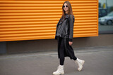Stylish beautiful pregnant girl walks along a city street.Wearing a black leather jacket, a black long dress and white high boots and black sunglasses.
