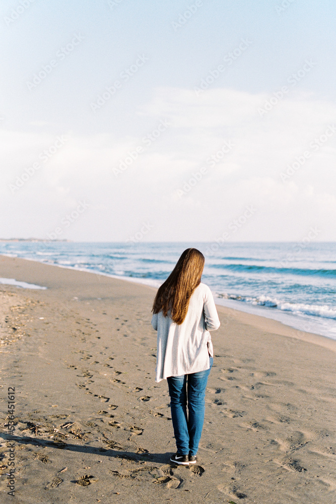Young woman in a long sweater walks along the sandy beach. Back view