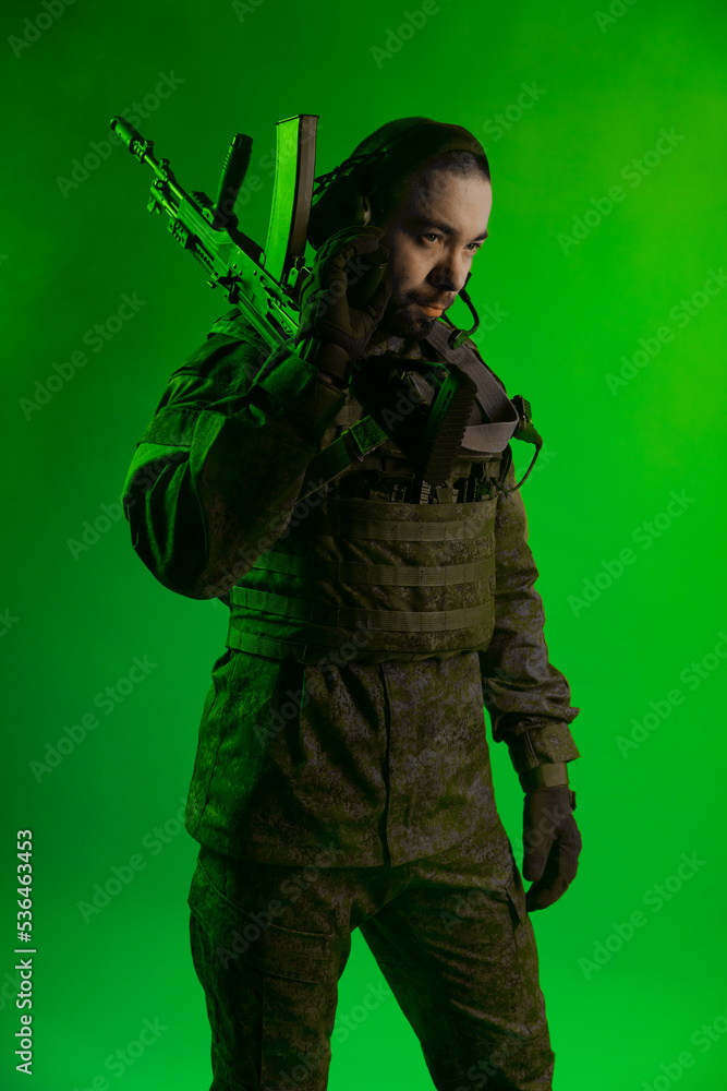 Fototapeta premium soldier in the studio on a green background with colored light. a man in military uniform with a gun, rifle or machine gun. military. airball player