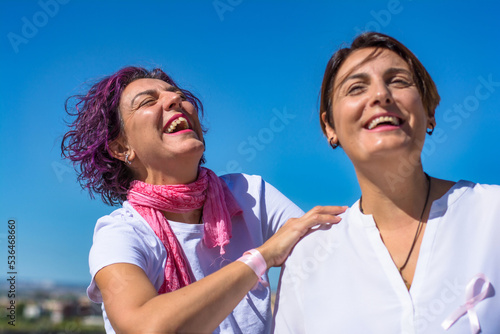 Breast Cancer Awareness Day Concept. Two smiling, happy women against a blue sky with pink scarf and and pink ribbon. Supreme concept. Pink October