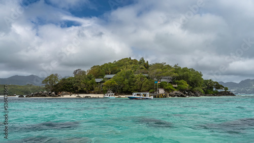 The island in the turquoise Indian Ocean is overgrown with tropical vegetation. Roofs of houses are visible through the foliage. The boats are moored at the pier. Boulders on a sandy beach.