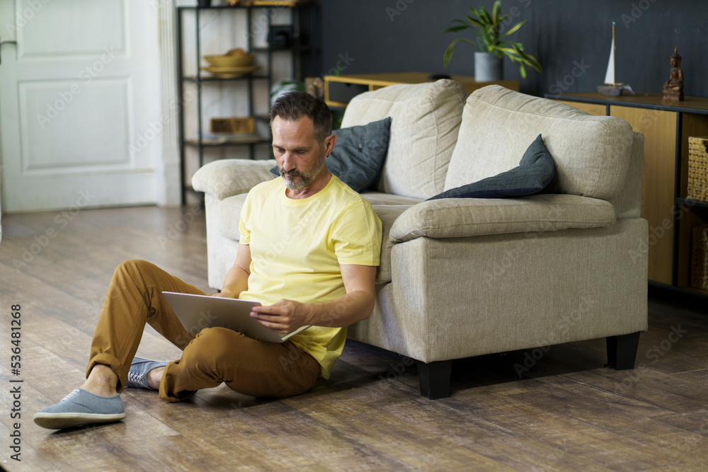 Man sitting on the floor with laptop computer in living room lean on sofa. Attractive mature man is browsing at his laptop, sitting at home near of sofa, wearing casual outfit.