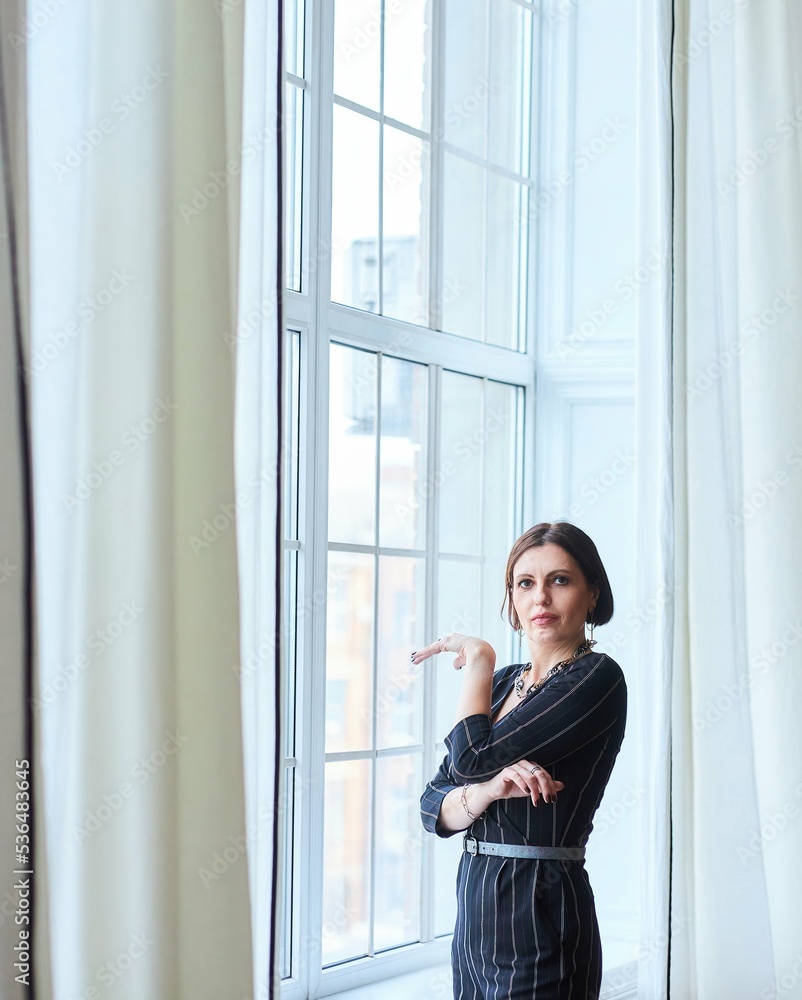 A stylish middle-aged woman in dark clothes stands at the window in a bright room. Style and fashion. A stylish middle-aged woman in dark clothes stands at the window in a bright room. Style and fashion.