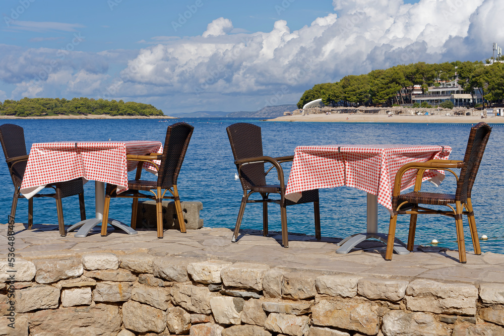 Heavenly place, set table of a restaurant on the Croatian Adriatic, on a stone wall, directly above the blue, crystal clear sea. In the background green overgrown islands and cloudy sky.