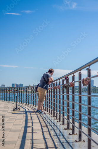 Fototapeta Naklejka Na Ścianę i Meble -  Children and a man walk across the bridge and look at the river. Various interesting things are attached to the railing. A hot summer day