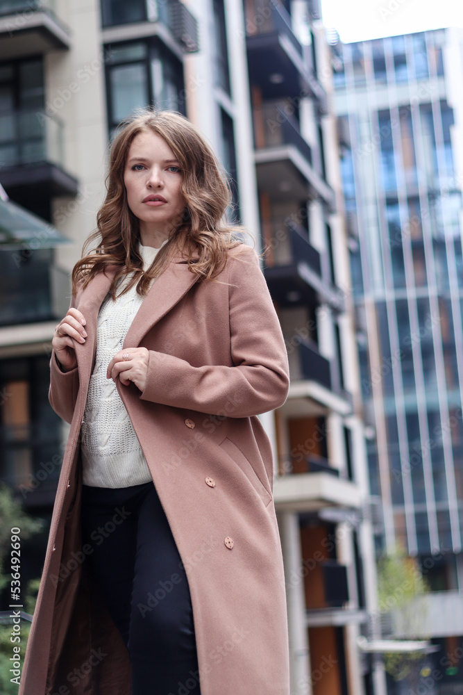 Street photo session of a girl with blond long hair in a beige coat. Photo shoot in the big city. Street photo session of a girl with blond long hair in a beige coat. Photo shoot in the big city.