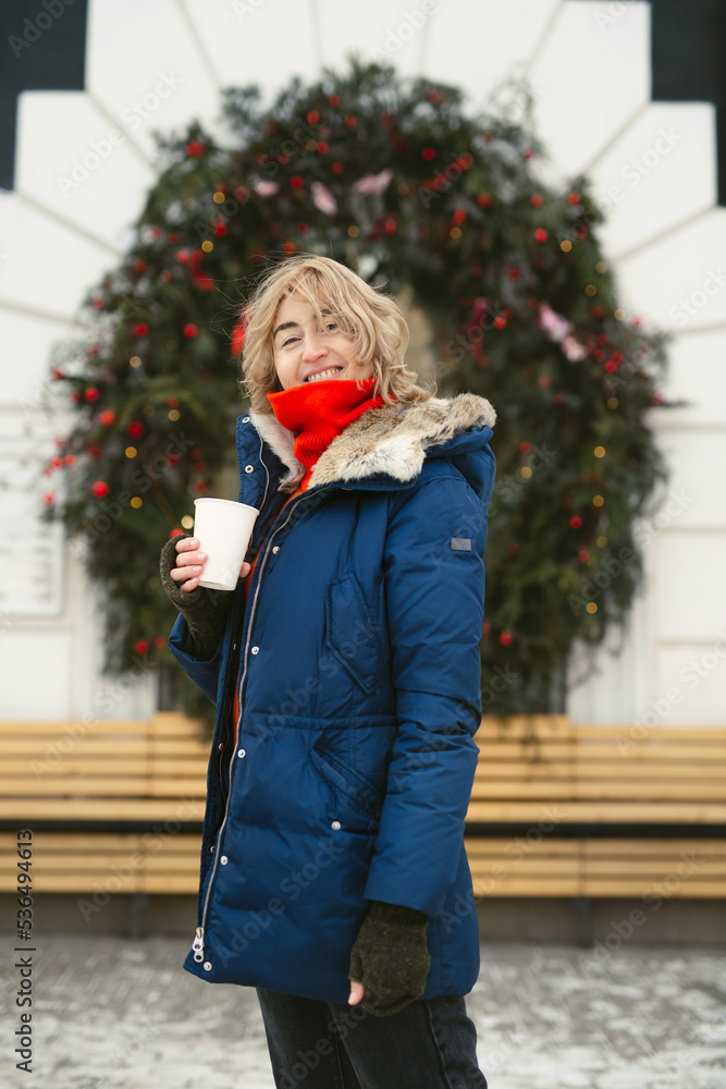 Woman drinks a hot drink on a city street against the background of a cafe window, which is decorated with a Christmas wreath. Woman drinks a hot drink on a city street against the background of a cafe window, which is decorated with a Christmas wreath.