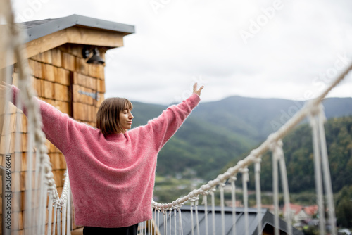 Fotografija Woman in pink sweater walks on rope bridge of wooden house highly in mountains