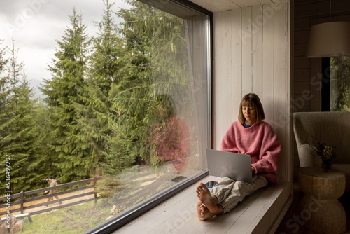 Woman works on laptop while sitting on a window with great view on mountains. Remote work from cozy place and escaping to nature concept photo