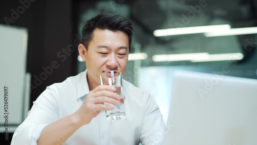 Asian employee man works in the modern office at the workplace sitting at the laptop computer drinking clean water from a glass. Business man, worker, quenches thirst Indoors