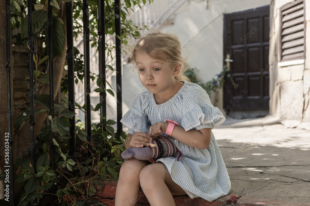 A little girl is sitting on the stone stairs and holding her toy in her ...