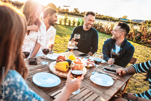 Group of friends having fun at bbq outside dinner in home garden - Happy people cheering red wine sitting outdoor at dining table - Social gathering, youth, food and beverage concept