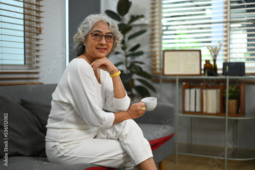 Happy elderly woman relaxing on couch in living room and drink herbal tea, enjoy leisure weekend at home