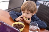 Cute little boy sit on chair at table and watch cartoon on tablet computer top view. Kid of kindergarten age eat chocolate balls with milk and drink water from cup. Ready breakfast, gadget addiction