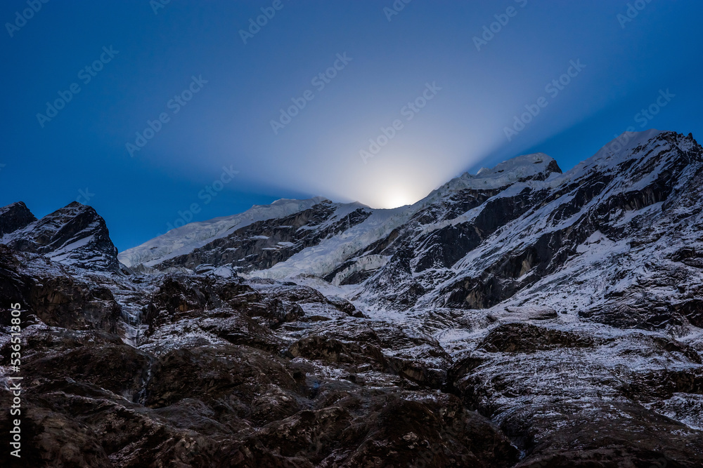 Light rays of the rising sun scatter behind the rocky peak and glacier in Himalayan mountains. Mountain landscape at high altitude in Nepal.