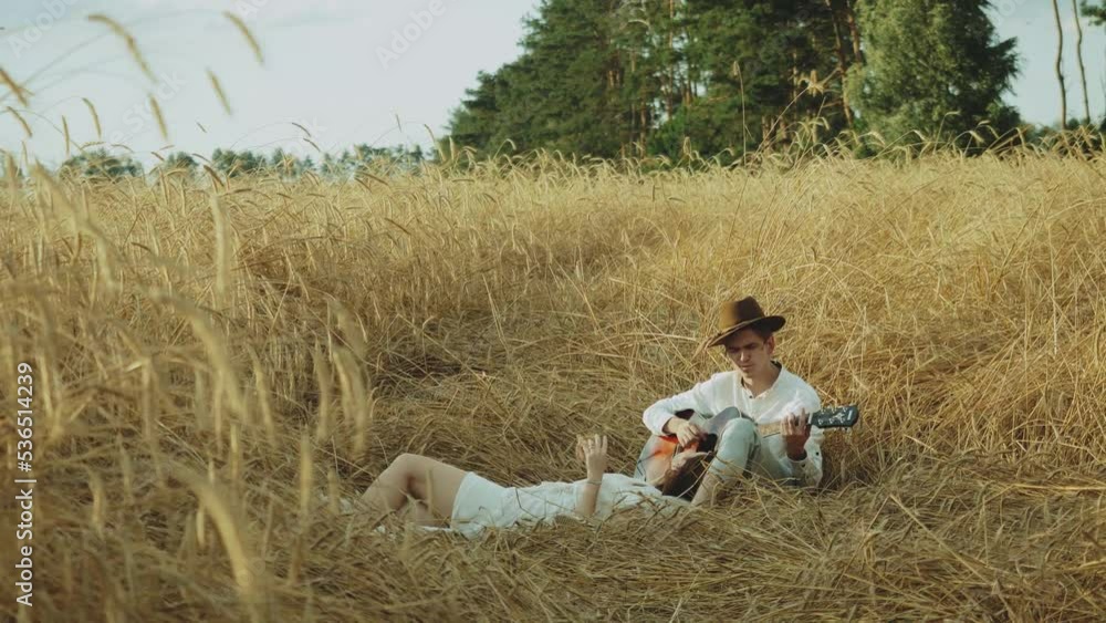 Attractive couple in the field of spikelets slow motion. Woman leans against the boy's back. Guy playing guitar girl listening. Attractive couple in the field of spikelets slow motion. Woman leans against the boy's back. Guy playing guitar girl listening.