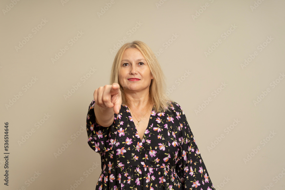 Pretty blond Caucasian woman in a dark patterned dress pointing happily at the camera, isolated over beige background. Pretty blond Caucasian woman in a dark patterned dress pointing happily at the camera, isolated over beige background.
