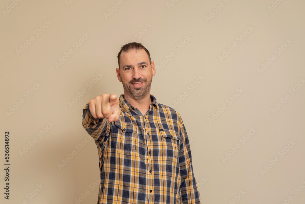 Hispanic man with a beard dressed in a plaid shirt pointing at the camera with his index finger, isolated on beige studio background. Hispanic man with a beard dressed in a plaid shirt pointing at the camera with his index finger, isolated on beige studio background.