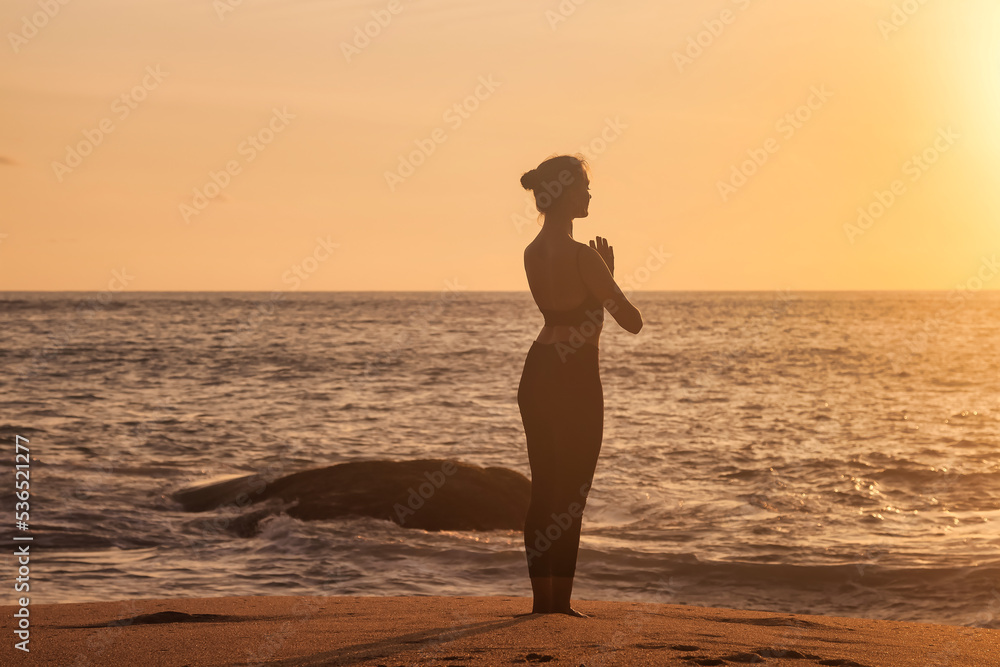 Rear view silhouette young woman does asana yoga position on tropical sea coast sandy beach, relaxing at sunset. Female performs exercises for healthy lifestyle to restore strength, spirit. Copy space