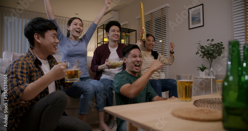 Group of young adult friend man and woman asia people sit at sofa couch joy night party fun game FIFA world cup live TV at home eat snack bowl drink beer bottle glass jump mad happy win exult face photo