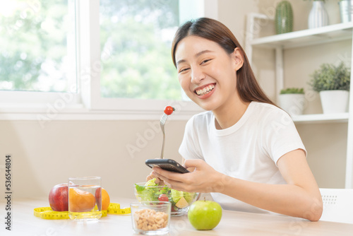 Dieting, smile asian young woman eating, holding fork at tomato, diet plan nutrition with fresh vegetables salad, enjoy meal while using smartphone. Nutritionist of healthy, nutrition of weight loss.