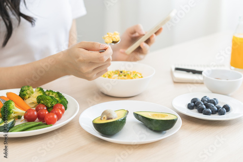 Dieting asian young woman eating keto food, hand holding spoon eating egg salad in bowl, diet plan nutrition with vegetables while using smartphone. Nutritionist of healthy, nutrition of weight loss.