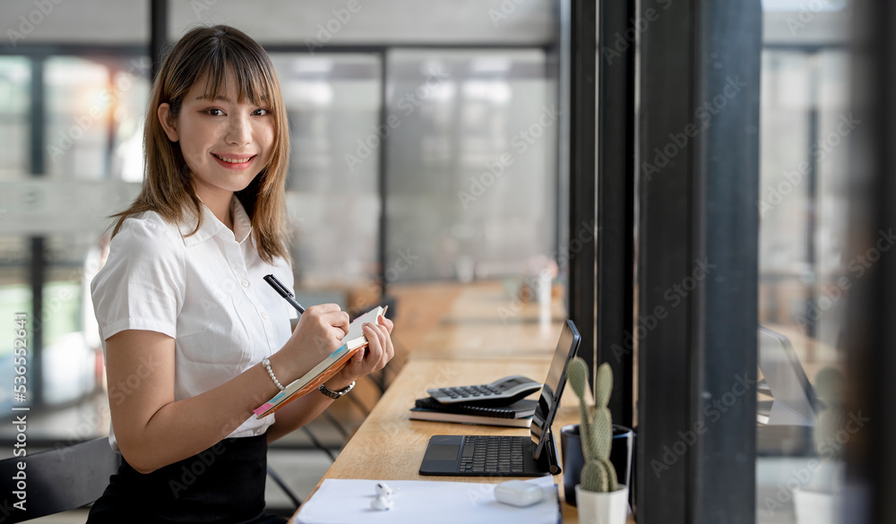 Obraz premium Charming Asian woman with a smile looking at camera while sitting and holding notebook at the office.