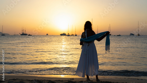 Rear view of a woman in a white dress, barefoot on the beach as she watches the sunset in Formentera, Spain