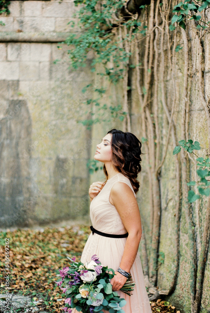 Bride with a bouquet stands near a stone wall entwined with ivy