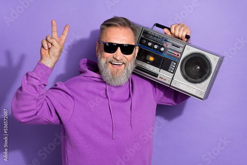 Portrait of handsome retired healthy man wear purple hoodie showing v-sign hold boombox on shoulder isolated on violet color background
