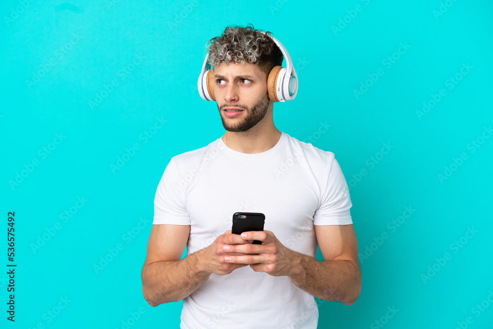 Young handsome caucasian man isolated on blue background listening music with a mobile and thinking Young handsome caucasian man isolated on blue background listening music with a mobile and thinking