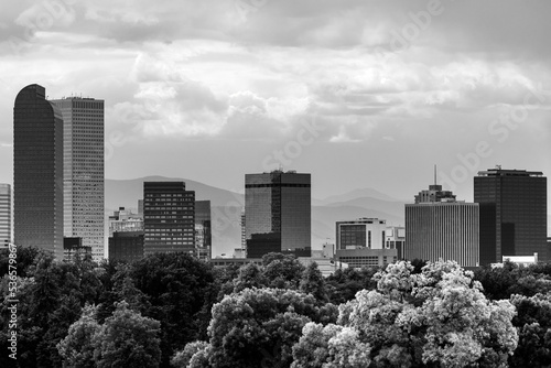 Black and white of the Denver Skyline with trees in front and mountains behind.