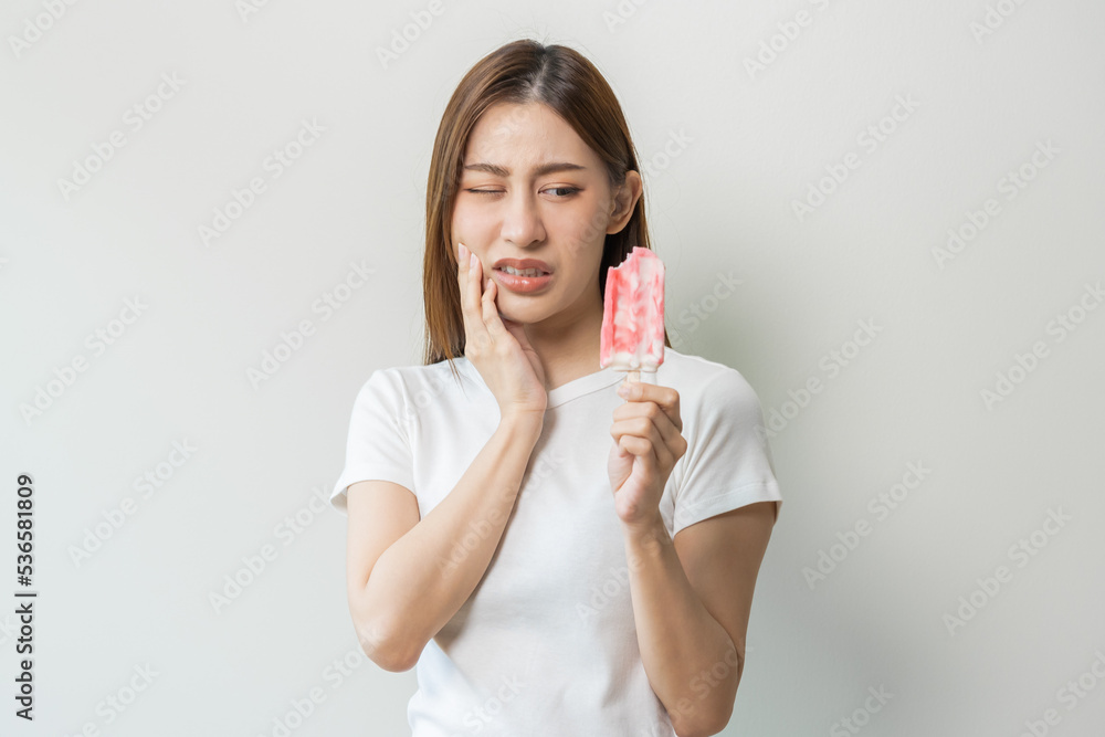 Face expression suffering from sensitive teeth and cold, asian young woman, girl hand touching her cheek, feeling hurt, pain eating ice cream, lolly. Toothache molar tooth at home, dental problem. Face expression suffering from sensitive teeth and cold, asian young woman, girl hand touching her cheek, feeling hurt, pain eating ice cream, lolly. Toothache molar tooth at home, dental problem.