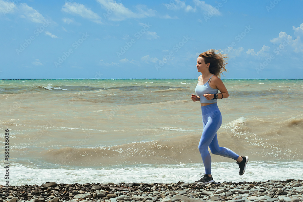 a young girl athlete with long wavy hair in a blue tracksuit runs along the seashore, on which a storm is raging against a blue sky on a sunny day