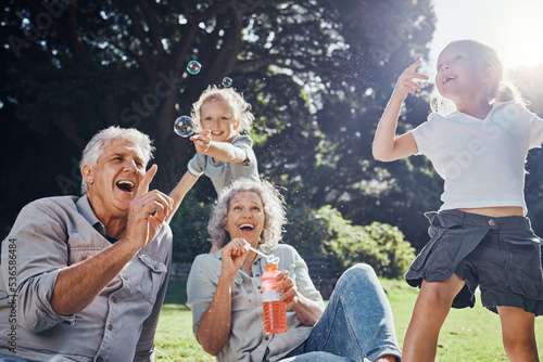 Grandparents, bubbles and children play in park happy together for fun, joy and outdoor happiness. Retired, smile and excited elderly senior couple, girl grandkids and love playing outside in nature photo