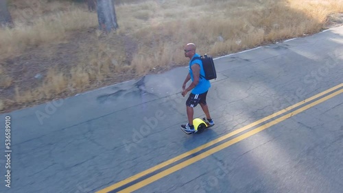 aerial footage of an African American man wearing a backpack riding a Onewheel electric skateboard at the Rose Bowl surrounded by dry brush and lush green trees, grass and plants in Pasadena photo